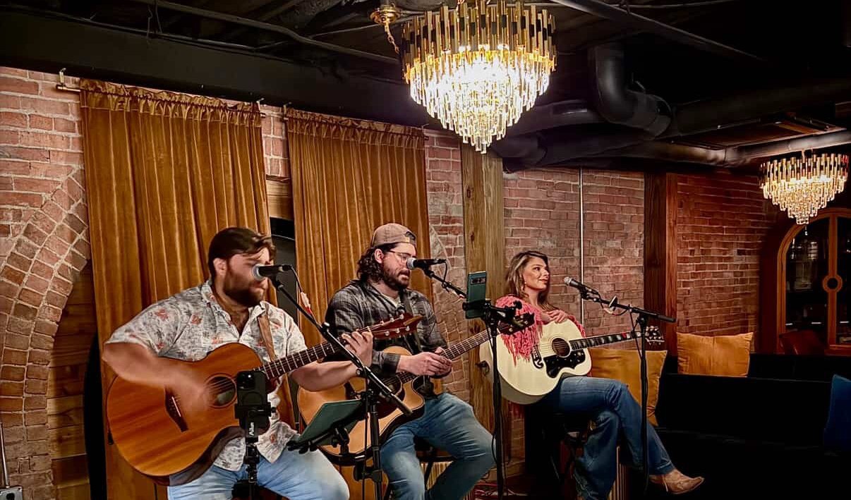 Matt Long performing in a songwriter round with two other guitarists in an upscale brick-walled room with chandeliers
