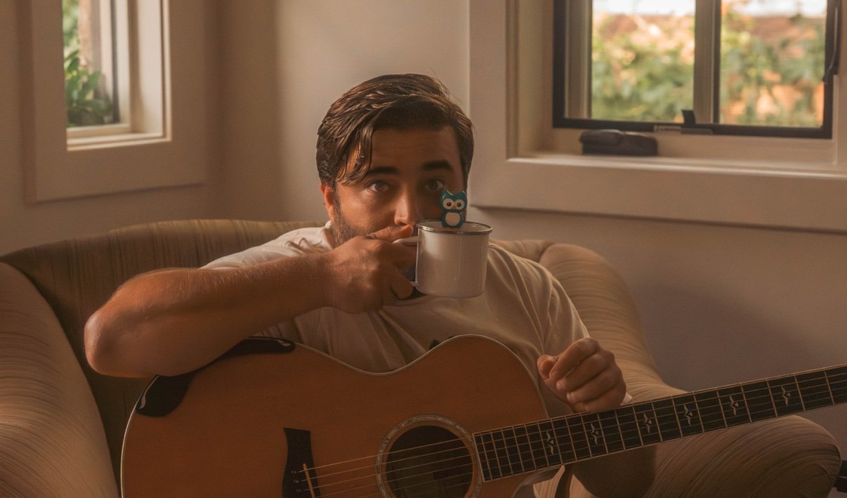 Matt Long relaxing in an armchair with his acoustic guitar across his lap, sipping from a mug with a little owl on it