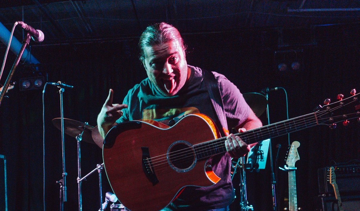 Matt Long grinning at the camera with his acoustic guitar on a club stage, a drum kit behind him