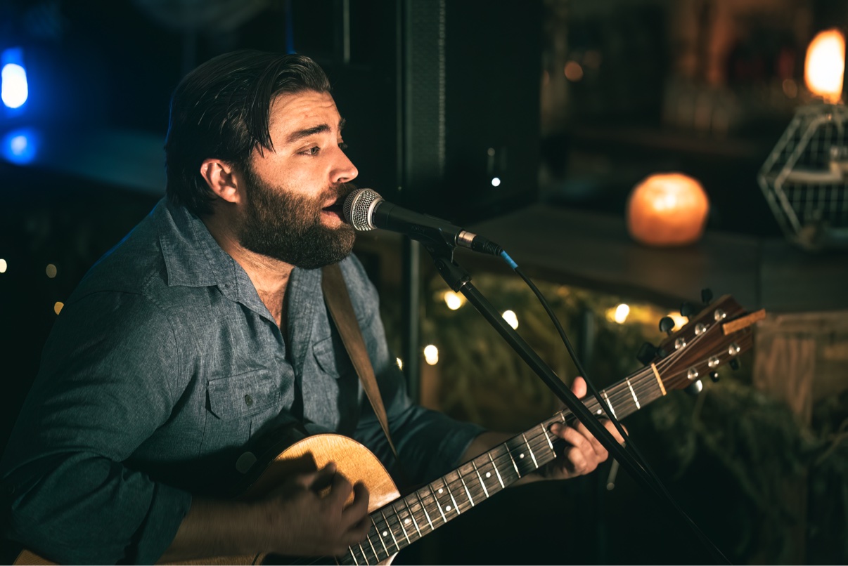 Close-up of Matt Long in a blue shirt singing into a microphone and playing acoustic guitar, with warm string lights and a glowing candle behind him at an intimate evening gathering