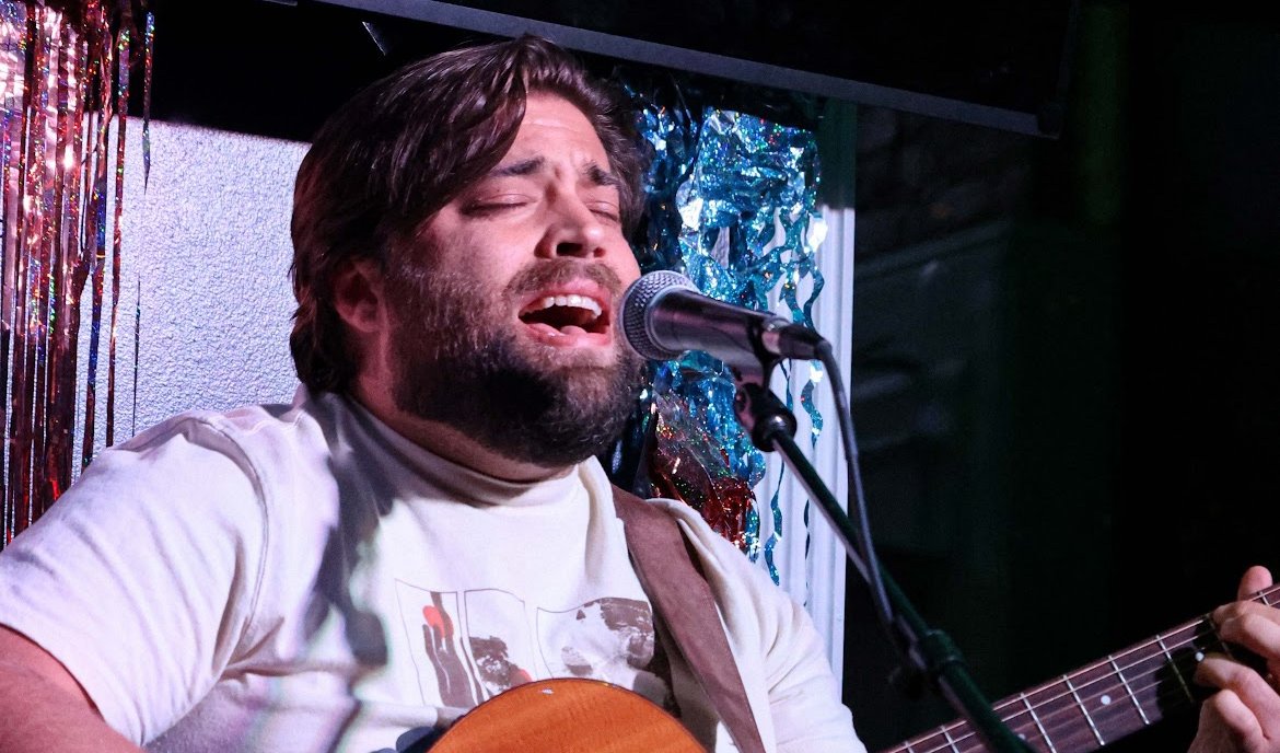 Close-up of Matt Long singing into a microphone and playing acoustic guitar on a dark club stage, with tinsel lights behind him