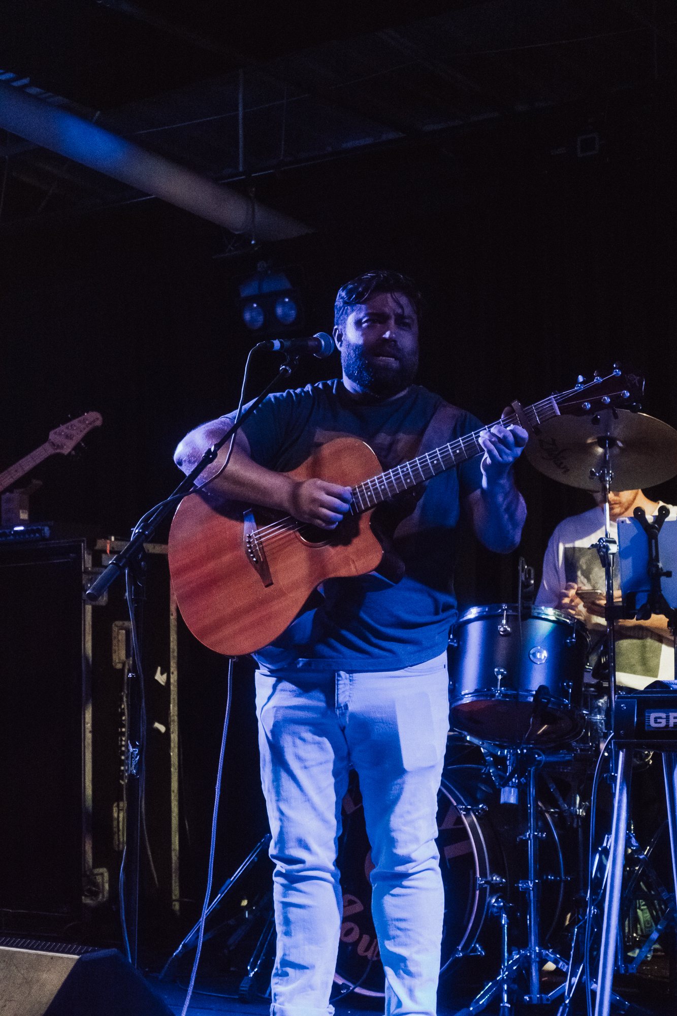 Matt Long playing acoustic guitar and singing on a blue-lit venue stage, with a drum kit behind him