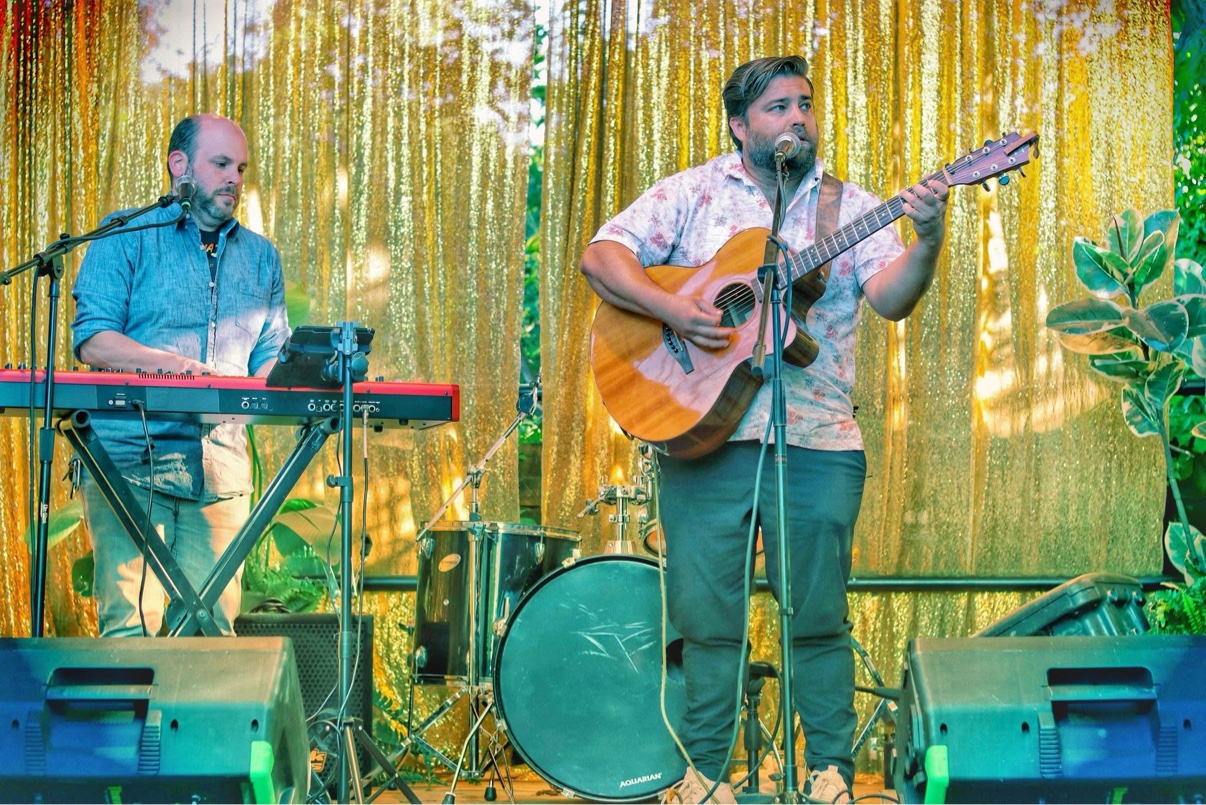 Matt Long singing and playing acoustic guitar alongside a keyboard player in front of a gold sequin curtain at an outdoor event, with drums between them