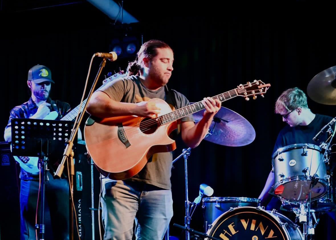 Matt Long playing acoustic guitar on stage with his full band, a bass player and drummer alongside him