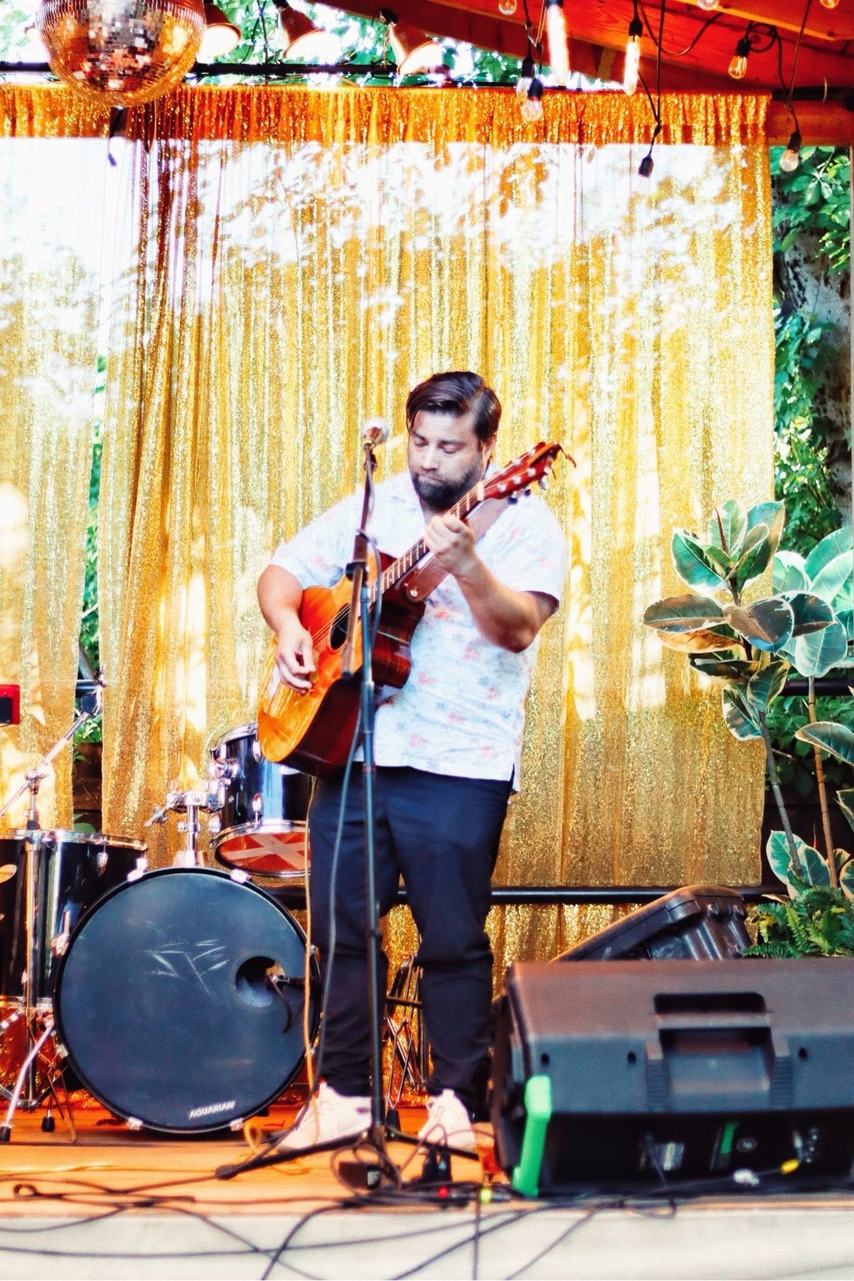 Matt Long looking down at his acoustic guitar as he plays in front of a gold sequin curtain, with string bulbs and a disco ball overhead
